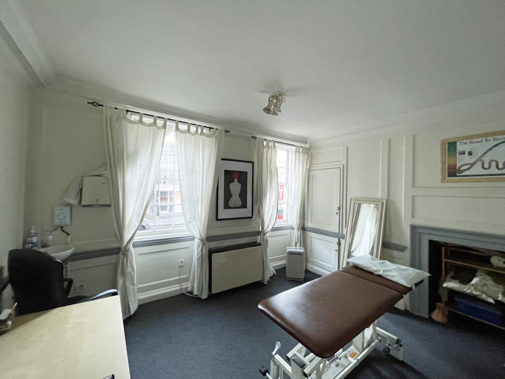 A clinic treatment room with tall ceilings, big windows and a brown bed in the centre of the room